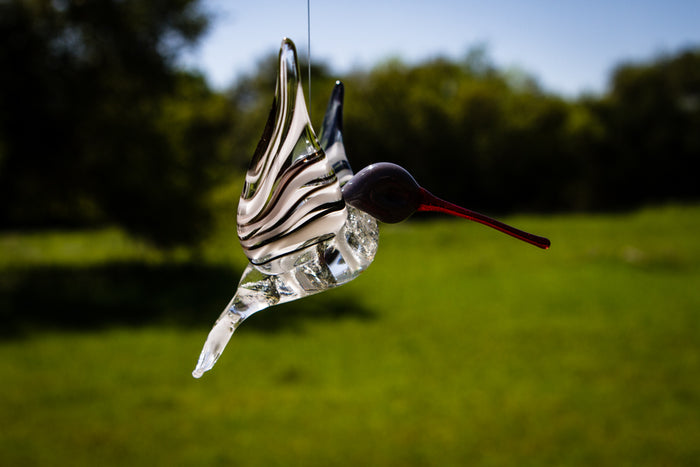 Glass hummingbird with black and white swirls and a red beak, hanging in daylight.