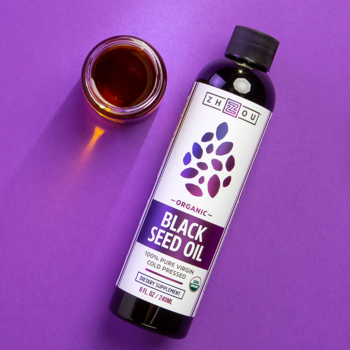 Zhou Nutritions Black Seed Oil bottle sits on a purple surface next to a small glass cup of the oil. The label highlights its 100% Pure Virgin, Cold Pressed quality as a dietary supplement, 8 fl oz/240ml.