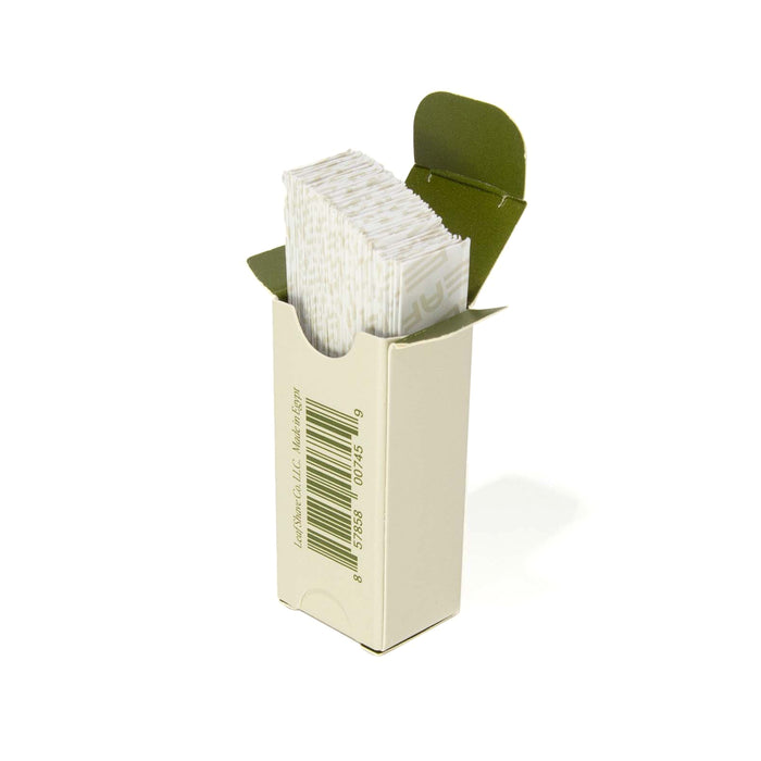 A beige cardboard box with a green interior lid, labeled as Leaf Shave Blade Refills, is partially open to reveal a stack of folded white strips inside. It contains stainless steel blades and features a barcode on the front.
