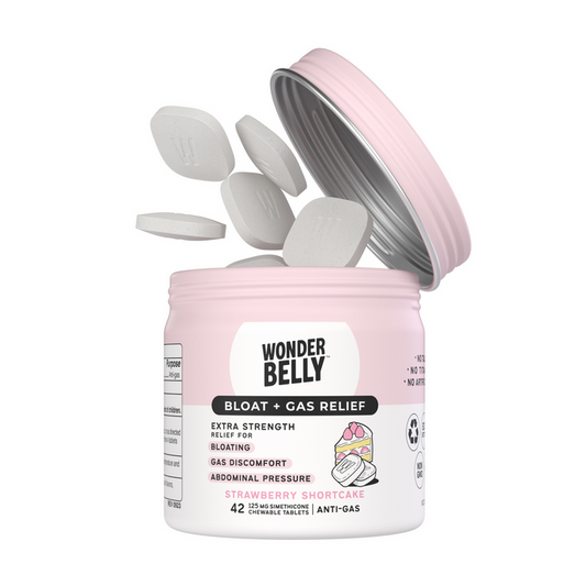 A pink and white container with Wonderbelly branding is open, revealing oval simethicone tablets labeled Bloat + Gas Relief with a strawberry shortcake flavor. Tablets float above the container, ready to offer quick relief from bloat and gas discomfort.