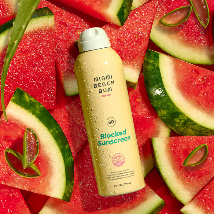 A can of Miami Beach Bum Blocked BOV Spray - SPF 50, a reef-friendly mineral sunscreen, sits on fresh watermelon slices. The yellow bottle glistens with water droplets against the vibrant backdrop of green rinds and red flesh.