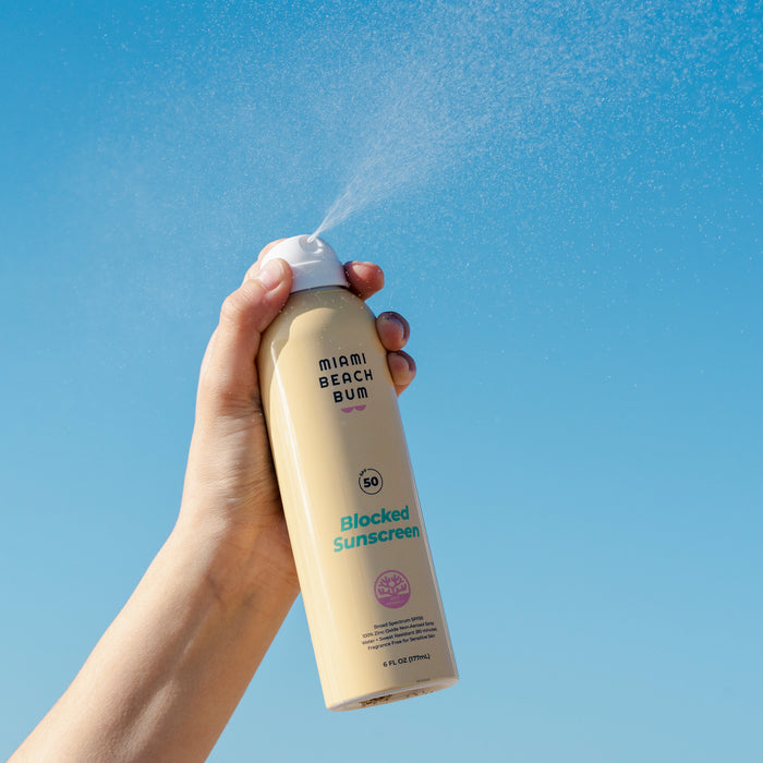 A hand sprays Miami Beach Bum’s Blocked BOV Spray SPF 50 against a clear blue sky, offering reef-friendly protection.