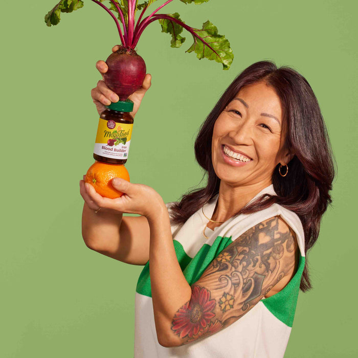A smiling woman with long hair and a floral tattoo holds a beetroot with its leaves atop a MegaFood Blood Builder bottle, balanced on an orange. Set against a light green background, the scene symbolizes the journey to naturally boost iron levels.