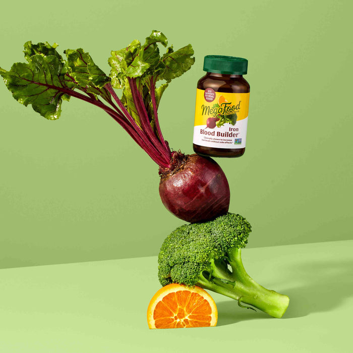 A MegaFood Blood Builder bottle sits on a beetroot, creatively balanced on broccoli and half an orange against a light green background. This vibrant arrangement showcases the natural artistry of nutrition while designed to boost energy levels.