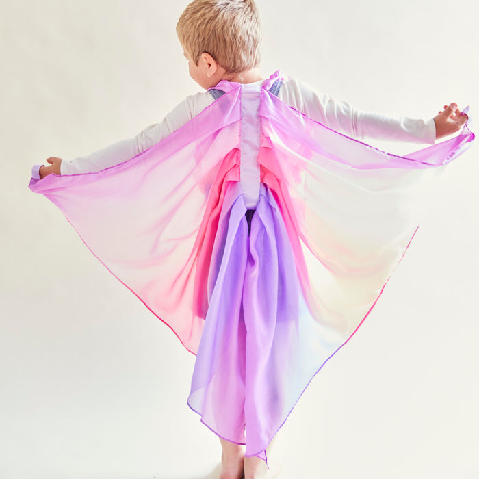 A blonde child is engrossed in dress-up, wearing the vibrant Blossom Fairy Wings from Sarahs Silks. They stand facing away, arms outstretched as if soaring, with pink, purple, and white mulberry silk against a light background.