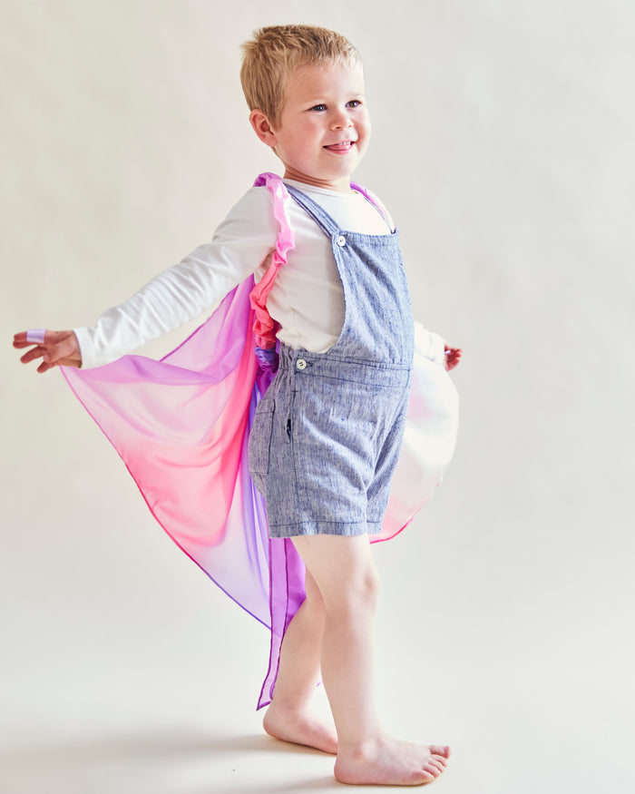 Barefoot and smiling, a young child in a white shirt and blue overalls embraces playful imagination by donning Sarahs Silks Blossom Fairy Wings, transforming their pink and purple cape into invisible wings that guide their whimsical adventures.