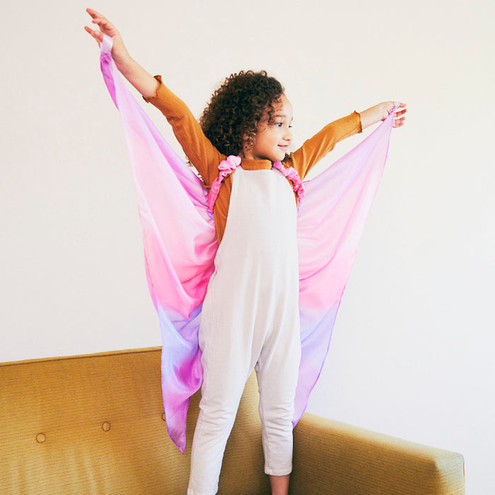 A child stands on a yellow couch, wearing Sarahs Silks Blossom Fairy Wings with pink and purple hues. Arms stretched joyfully, the smiling child gazes upward, embodying the enchantment of dress-up play against a simple light-toned backdrop.
