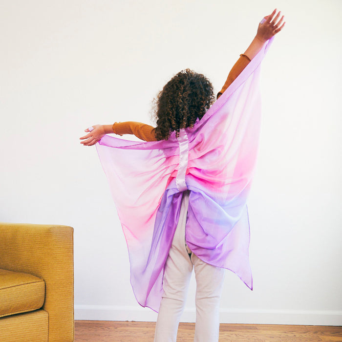 A child with curly hair, arms outstretched, wears Sarahs Silks Blossom Fairy Wings in pink, purple, and white. Engaged in dress-up play, the little one faces away with one wing longer than the other. A yellow couch is partially visible to the left.