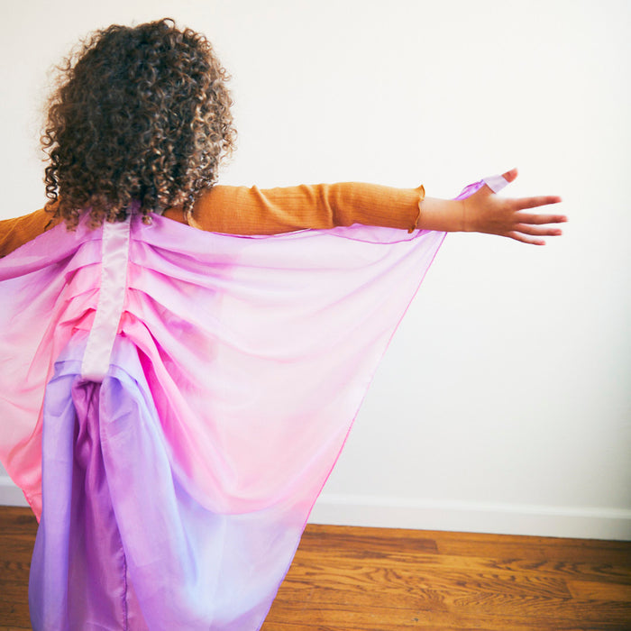 A curly-haired child, engaged in dress-up play, wears Sarahs Silks Blossom Fairy Wings in pink and purple mulberry silk. Standing on a wooden floor against a white wall, they face away from the camera with arms outstretched.