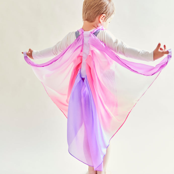 A child with short hair in a white top spreads out Sarahs Silks Blossom Fairy Wings, showcasing their vibrant pink and purple colors. These mulberry silk wings flow elegantly, inspiring an imaginative dress-up play scene on a light background.