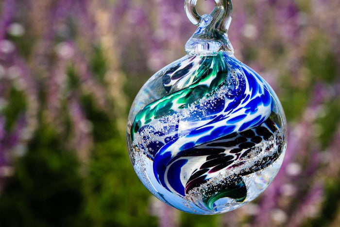 Blue green glass cremation orb with swirling multicolor pattern hanging outdoors against a garden backdrop.