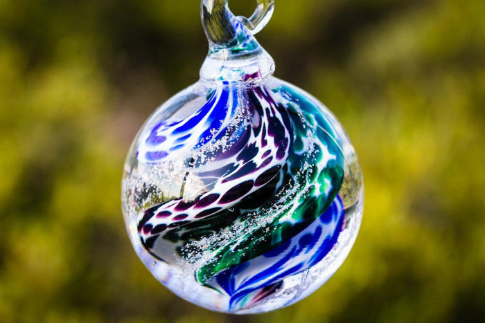 Close up of a multicolored swirled glass cremation orb in blue green and purple tones, hanging against a blurred garden background.