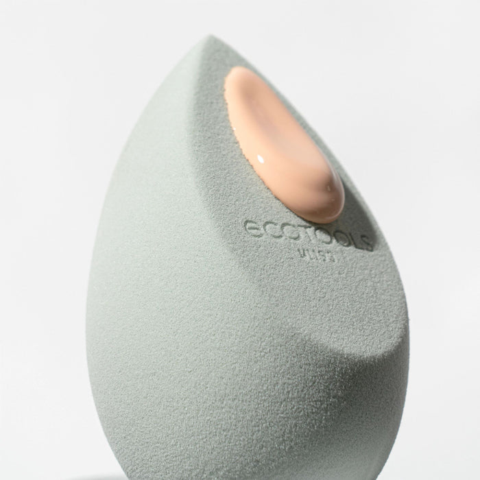 A close-up of the EcoTools Blurring Blender Duo shows a drop of beige foundation on the textured, cruelty-free sponge, highlighting its commitment to ethical beauty against a plain, light background.