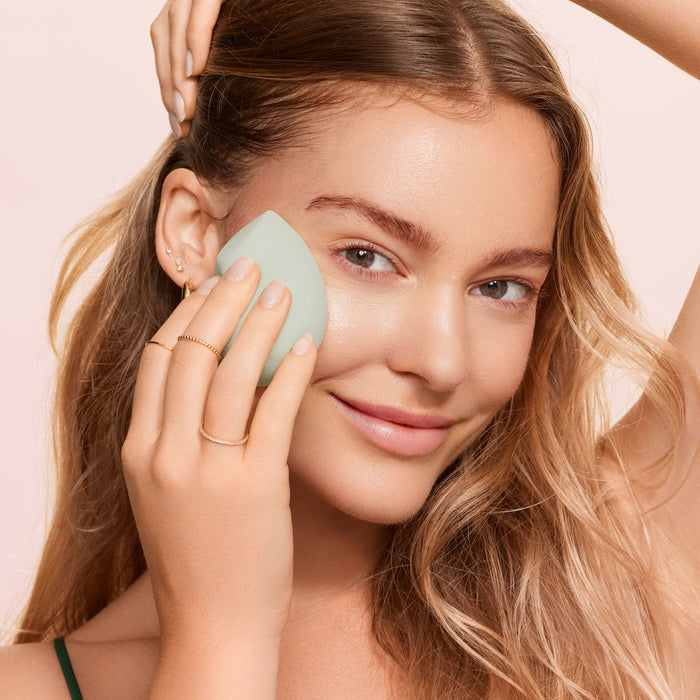 A woman with long blonde hair smiles while applying the EcoTools Blurring Blender Duo in pale green to her left cheek. Shes wearing a thin-strap top and multiple earrings, with simple rings on her fingers against a soft, pale background.
