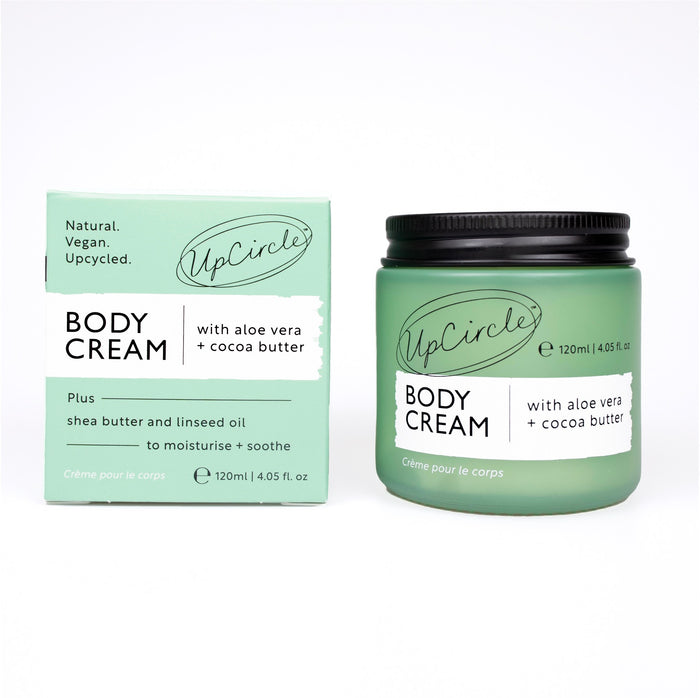 An UpCircle Body Cream with Aloe Vera + Cocoa Butter is shown in a green jar with a black lid beside a matching box, featuring text about key ingredients: aloe vera, cocoa butter, shea butter, linseed oil, and nourishing date seed.
