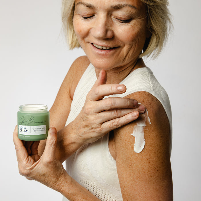 A person with short hair smiles while applying UpCircle Body Cream with Aloe Vera + Cocoa Butter, holding its jar adorned in green packaging with white text. Clad in a sleeveless white top, they apply it to their upper arm against a plain background.