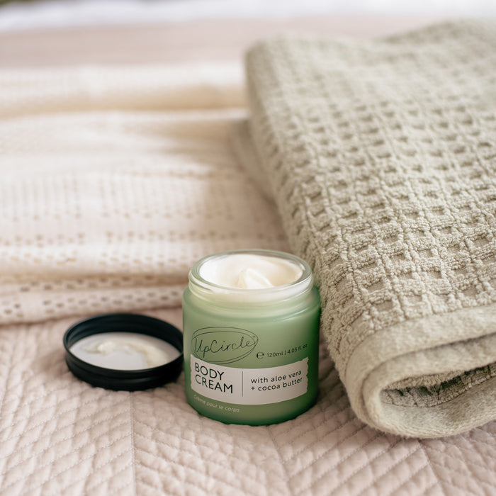 An open jar of UpCircle Body Cream with Aloe Vera + Cocoa Butter rests on a soft beige quilt. The green jars black lid lies nearby, alongside an elegantly rolled textured beige towel, enhancing the soothing scene.