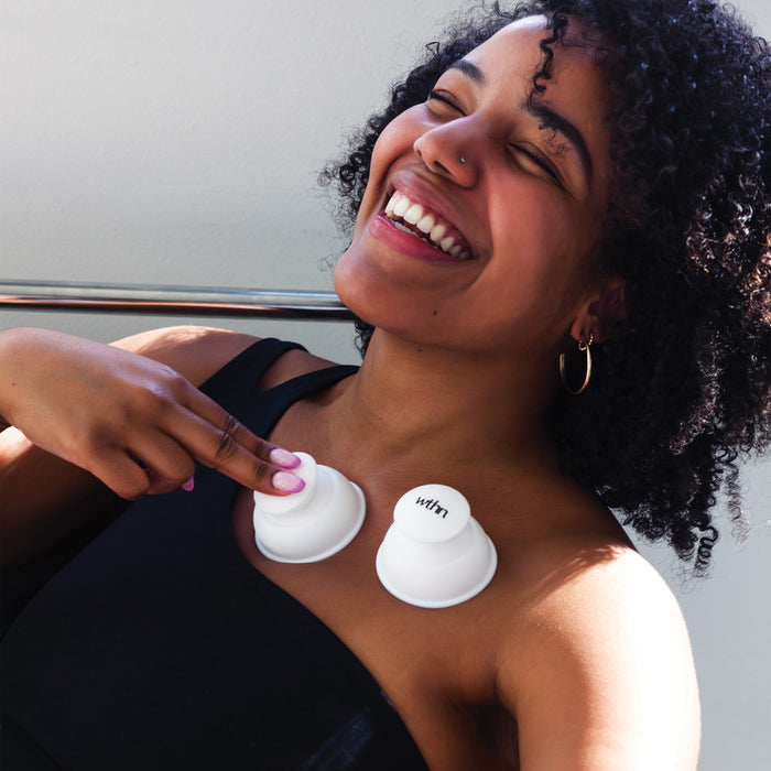 A smiling woman with curly hair uses the WTHN Body Cupping Kits two white silicone cups on her chest. Dressed in a black top and hoop earrings, she enjoys the soothing techniques of Traditional Chinese Medicine.