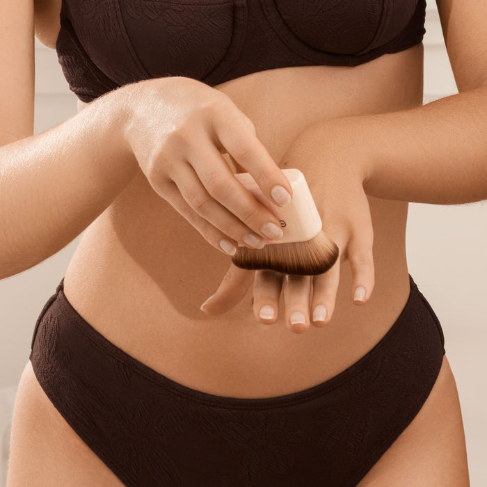Wearing dark underwear, a person gently uses the EcoTools Body + Face Self-Tan Blending brush on their hand for a streak-free finish. This cruelty-free, vegan tool ensures flawless application against a neutral backdrop.