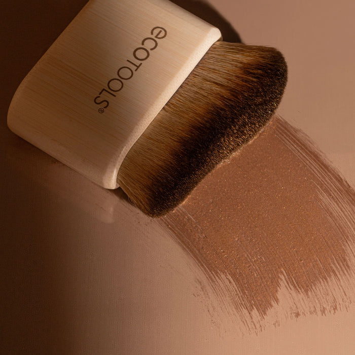 EcoTools Body + Face Self-Tan Blending brush, with soft bristles sweeps a streak-free stroke of shimmery brown powder across a smooth surface. Known for its cruelty-free, vegan quality, it features a light-colored handle.