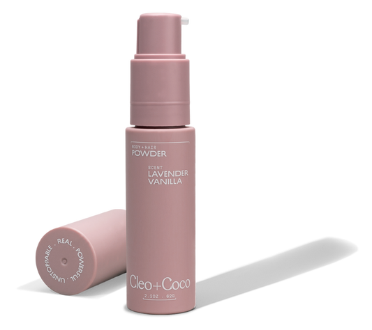 A pink bottle of Cleo & Coco Body + Hair Powder stands upright with its cap beside it. The label and cap display white text, highlighting this aluminum-free formula made from natural ingredients including arrowroot powder.