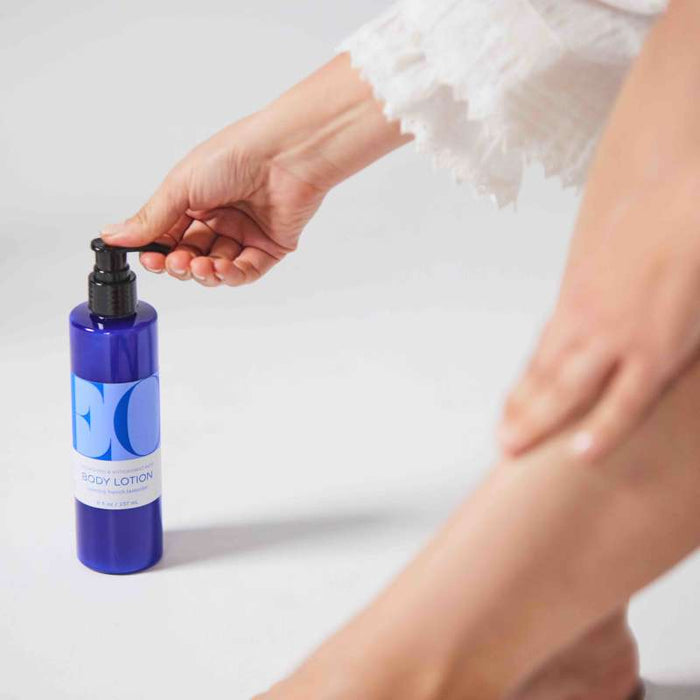 In a bright, minimalistic setting, someone in white pumps lotion from a blue EO Products bottle labeled Body Lotion as they moisturize their legs.