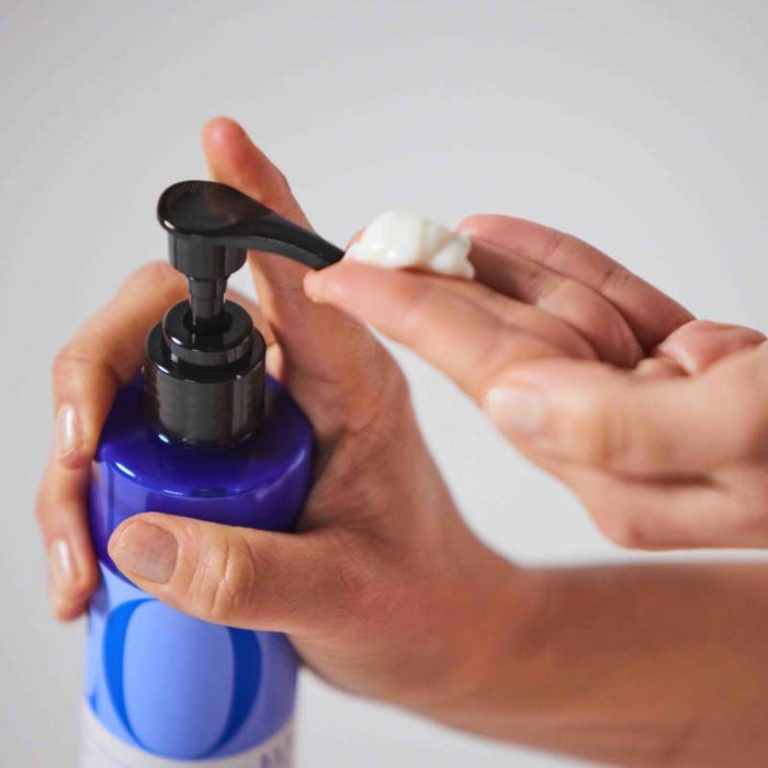 An individual applies EO Products Body Lotion, enhanced with jojoba oil, from a blue pump bottle onto their fingers. The hand waits under the nozzle for the nourishing blend, set against a soft gray backdrop that highlights the scenes elegance.