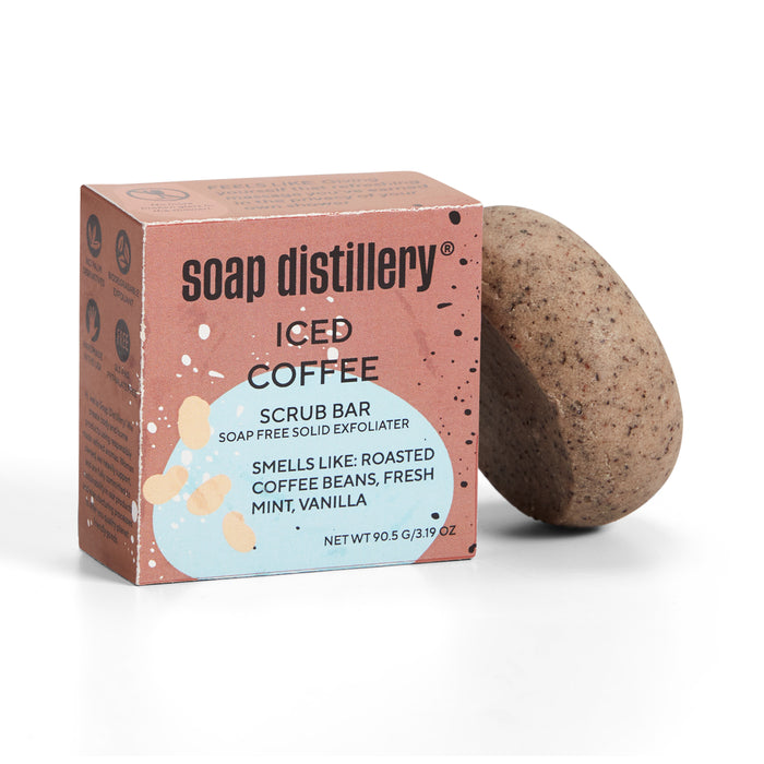 A rectangular pink box labeled Soap Distillery Body Scrub Bar sits beside a speckled, oval exfoliating soap bar. This plastic-free product features scents of roasted coffee, mint, and vanilla while moisturizing your skin.
