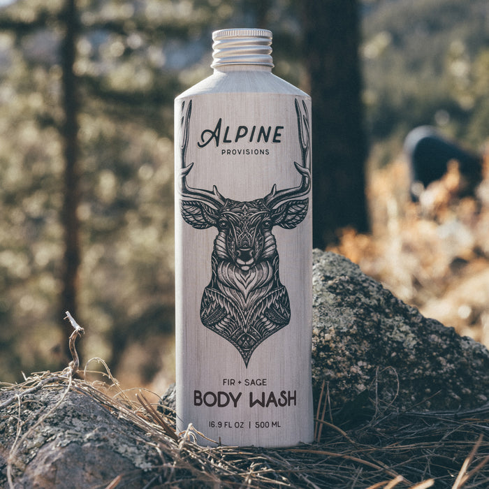 A 16.9 fl oz Alpine Provisions body wash with fir and sage scent, featuring a deer drawing, stands on a rock outdoors.