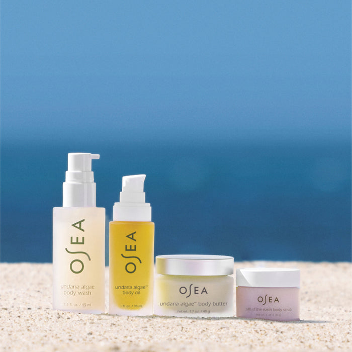 OSEAs Bodycare Bestsellers Set, infused with Undaria Algae, features a body wash, body oil, body butter, and scrub. Displayed on sand against a clear blue sky and ocean backdrop, these items in minimalist packaging are perfect for your travel set.