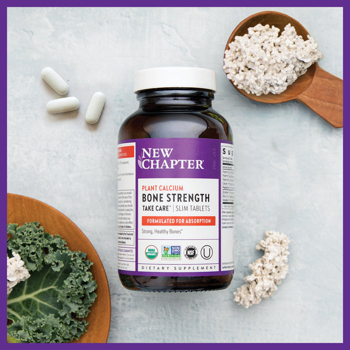 New Chapters Bone Strength Take Care™ bottle, featuring organic plant calcium for strong healthy bones, sits on a light surface with kale and calcium powder scoops. The label highlights absorption benefits, accompanied by three tablets beside the bottle.