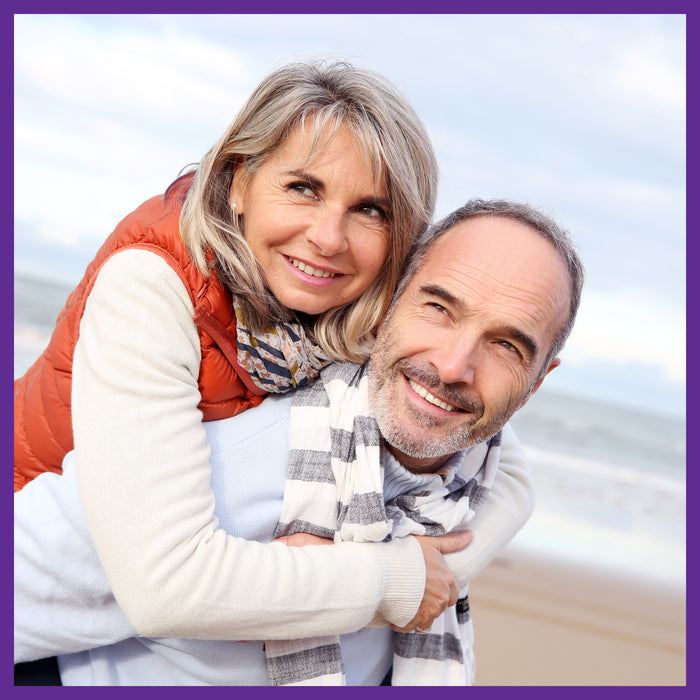 On a sandy beach with the sea as a backdrop, a gray-haired woman in a red vest and scarf joyfully hugs a man in a light blue sweater. They both radiate vitality thanks to New Chapters Bone Strength Take Care™ Organic Plant Calcium for strong, healthy bones.