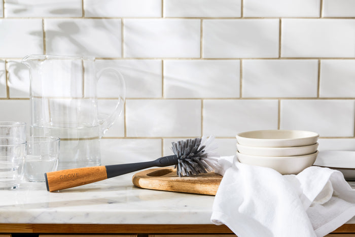A kitchen counter features a white tile backsplash, showcasing a clear pitcher, glasses, and a Grove Co. Bottle Brush Replacement Head on a wooden tray. Nearby are three white bowls stacked next to a universal handle wooden dish brush. Sunlight softly casts shadows across the scene.