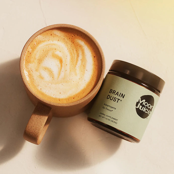 A frothy latte in a ceramic mug rests beside an open jar of Moon Juices Brain Dust—Adaptogens for Mental Stamina, Alertness & Concentration. The jar is tilted on a light surface, enveloped in soft warm light.