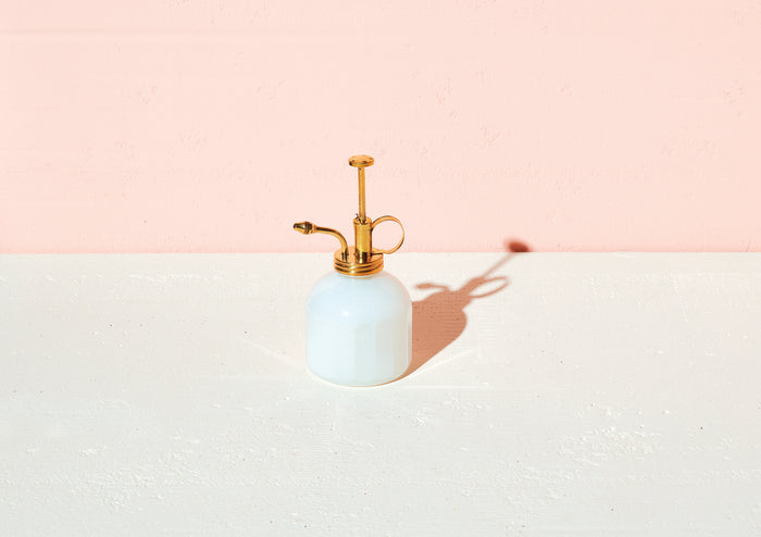 The Modern Sprout Brass & Glass Mister, a small white bottle with a brass top and pump, sits elegantly on a white surface. Ideal for indoor gardening, it stands against a pale pink wall with its shadow casting charmingly to the right.