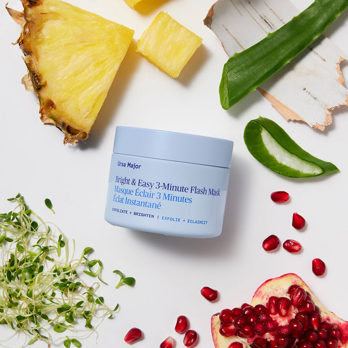 A blue jar of Ursa Major Bright and Easy Flash Mask is displayed on a white background, surrounded by pineapple, aloe vera, pomegranate seeds, bark, and green sprouts for a natural skincare vibe.