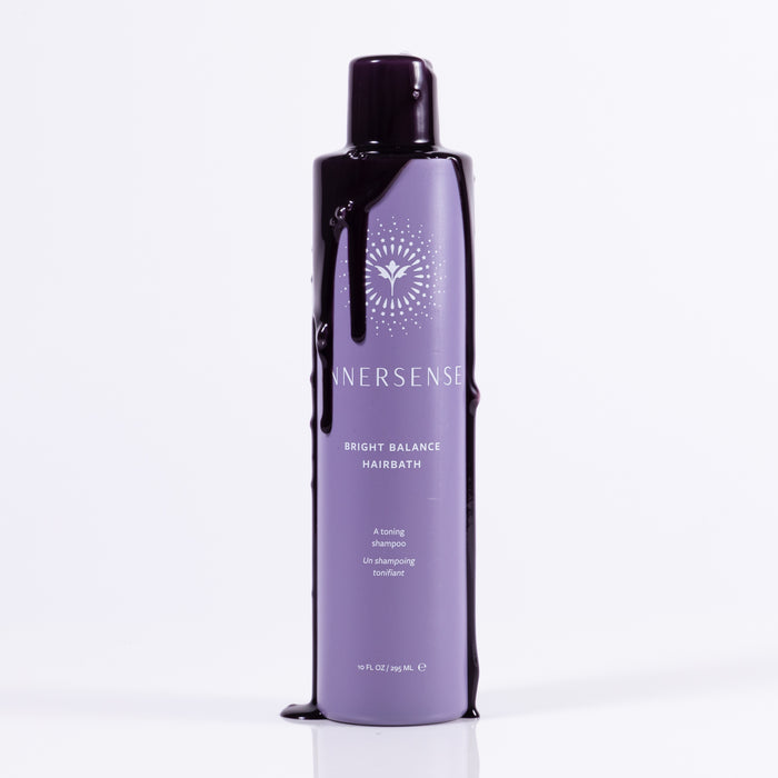 A bottle of Innersense Organic Beautys Bright Balance Hairbath, a vegan violet shampoo, stands upright with a sleek design featuring purple accents and dramatic dark liquid drips against a white background.