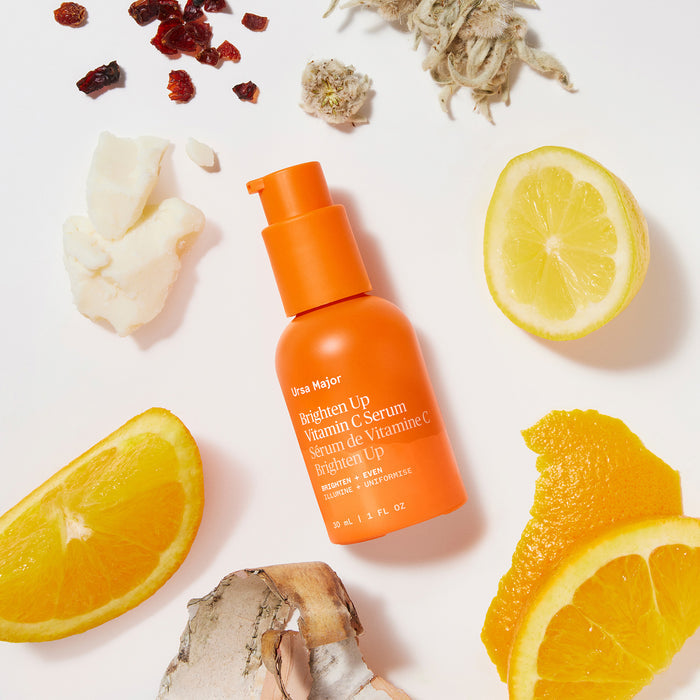An orange bottle of Ursa Major Brighten Up Vitamin C Serum sits on a white background, surrounded by lemon and orange slices, dried herbs, flower petals, and small chunks of a white substance.