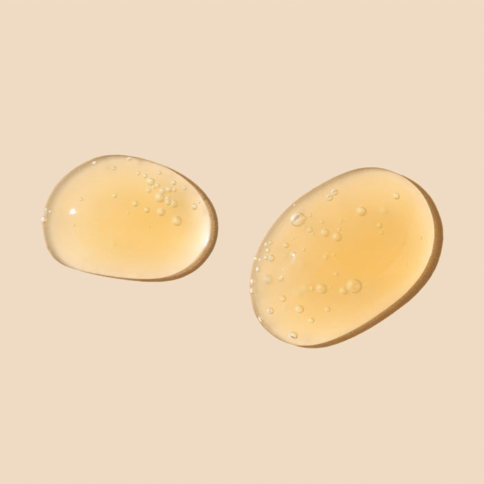 Against a beige backdrop, two drops of Wildcrafts Brighten Vitamin C Face Serum, translucent and amber with small air bubbles, promise a glossy, smooth texture designed to brighten skin and enhance its natural radiance.