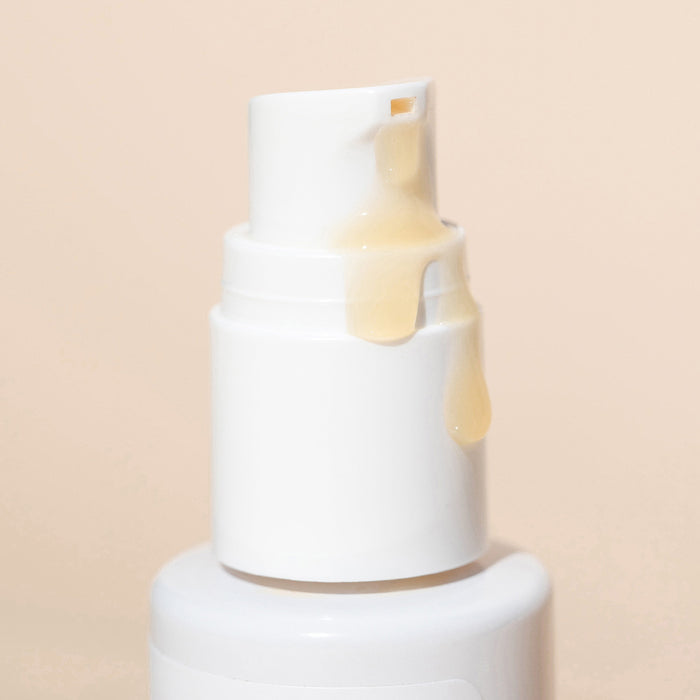 A close-up of a white pump bottle from Wildcraft dispensing Brighten Vitamin C Face Serum, revealing the thick, translucent cream enriched with hyaluronic acid as it flows down the nozzle against a beige background.