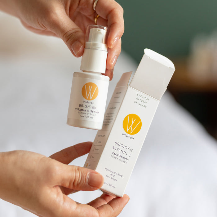 A person holds a Wildcraft Brighten Vitamin C Face Serum, featuring hyaluronic acid, with the white bottle and yellow logo contrasting against a blurred background, creating a cozy atmosphere ideal for brightening skin.
