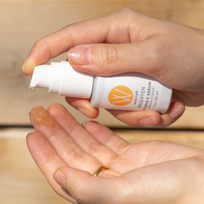 A person dispenses a small amount of Brighten Vitamin C Face Serum from a sleek white bottle with a pump. The label features an orange circle with a W for Wildcraft. Against the gentle glow of a wooden background, it promises to brighten skin for a radiant look.