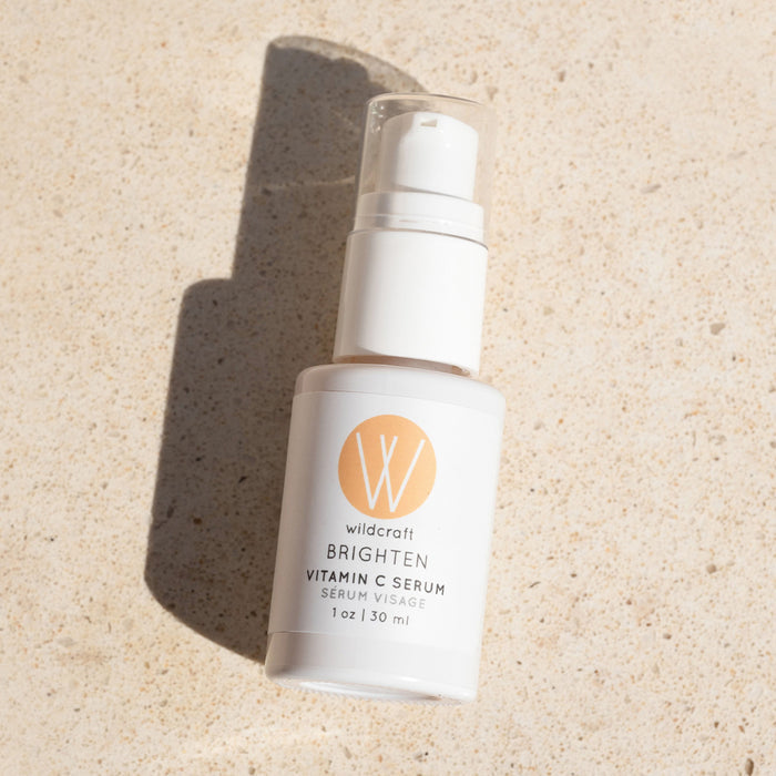 A Wildcraft Brighten Vitamin C Face Serum bottle stands on a stone surface, casting a shadow. The white label features an orange circle and black text. With hyaluronic acid and a clear pump dispenser, it provides a convenient daily skin-brightening solution.