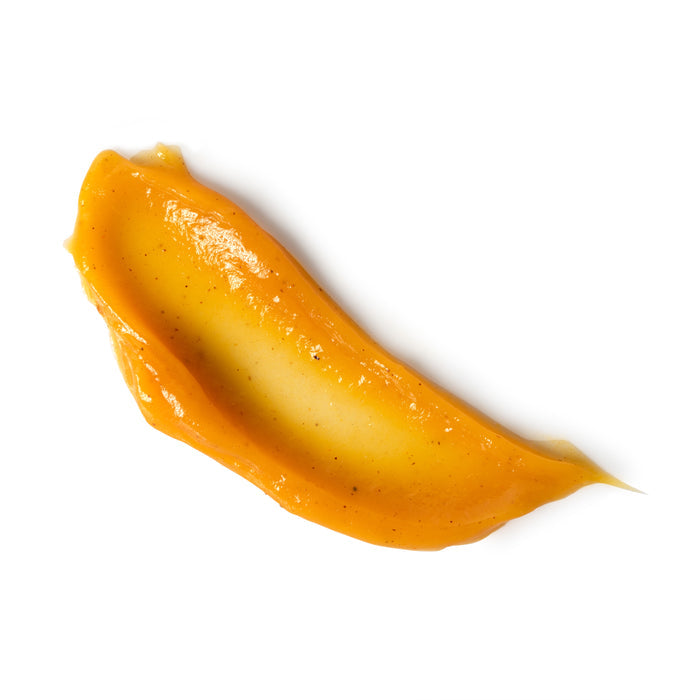 A smear of glossy, golden-yellow Brightening Pumpkin Honey Glycolic Mask from Andalou Naturals, infused with vitamin C, boasts a smooth texture and slight flecks as it elegantly spreads on a white background.