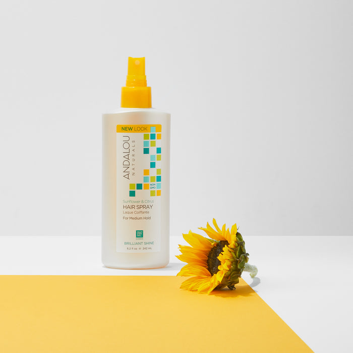 A bottle of Andalou Naturals Brilliant Shine Sunflower & Citrus Hair Spray, with vitamin C and fruit stem cell science, rests on a white surface beside a sunflower. It features a colorful square pattern and a yellow label for medium hold with a new look.