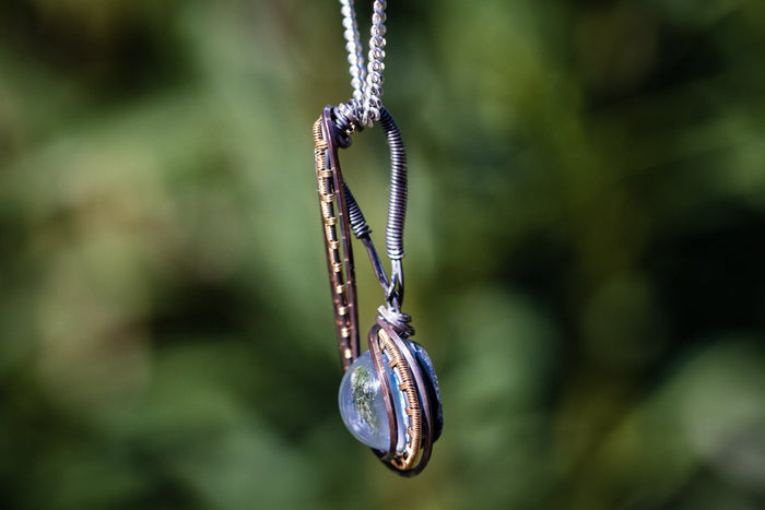 Close up of a Brizo pendant showing copper wire wrap around a blue glass cabochon on a silver chain.
