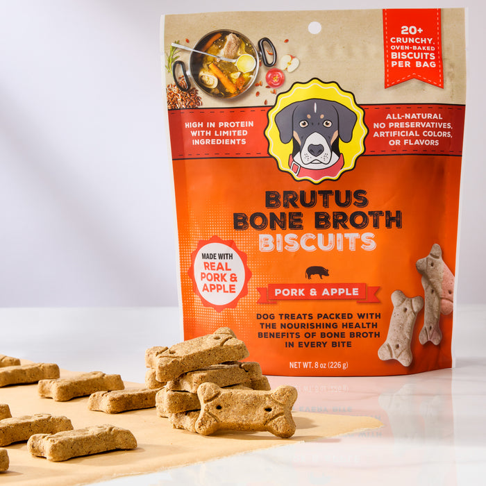 A Brutus Bone Broth Biscuits bag is surrounded by bone-shaped dog treats on wood. The orange packaging features a dog and highlights real pork, apple, and nourishing bone broth to promote joint health for your furry friend.