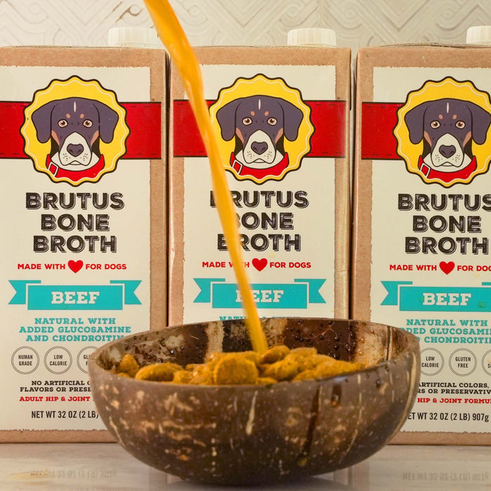 Three cartons of Brutus Bone Broth Hip & Joint by Brutus Broth, labeled Beef with a cartoon dog, are in the background. This all-natural broth supports hip and joint health and is poured into a dog food-filled bowl in the foreground.