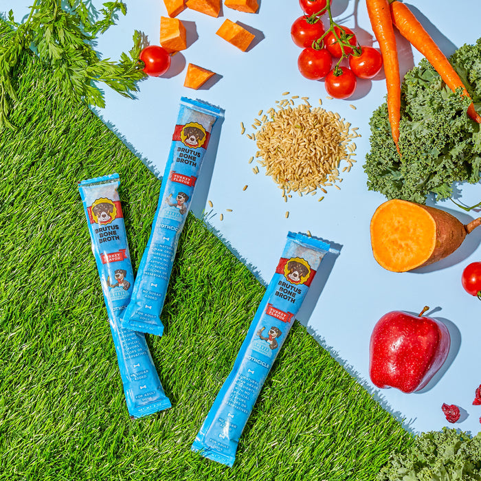 Three blue packets of Brutus Brothcicles by Brutus Broth rest on grass, surrounded by superfood ingredients: sweet potatoes, kale, carrots, cherry tomatoes, an apple, and rice—with a vivid blue background.