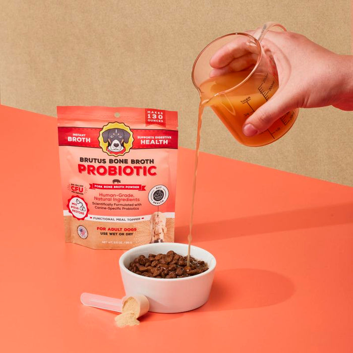 A hand pours liquid from a measuring cup into a bowl of dog food using Brutus on the Go: Probiotics Instant Bone Broth Mega Pack, enhancing gut health. A scoop filled with powder is ready to boost the benefits of bone broth for dogs, courtesy of Brutus Broth.
